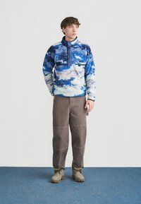 Noor mees seisab sinisel vaibal, kandes sinise mustriga fliisist pusa, pruune pükse ja beeže saapaid, vaadates küljele.