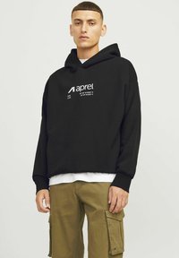 Mladi moški, oblečen v črno kapuco z belim napisom in logotipi, belo spodnjo majico in olivno zelene cargo hlače, stoji pred enobarvnim ozadjem.