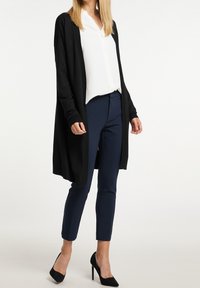 Žena v dlouhém černém cardiganu přes bílou košili s knoflíky, tmavě modré úzké kalhoty a černé boty na vysokém podpatku, stojící na bílém pozadí.