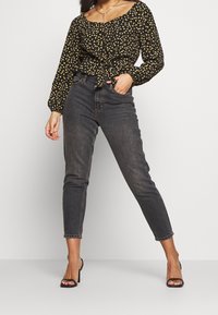 Kobieta w czarnej, kwiecistej bluzce z długim rękawem, w przetarciach na wysokim stanie czarnych dżinsach oraz czarnych szpilkach z odkrytymi palcami, pozująca z jedną ręką w kieszeni.