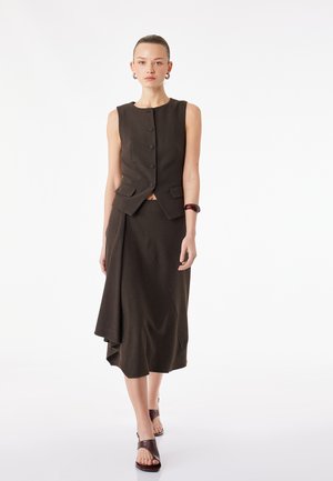 Kobieta idąca, ubrana w brązową bezrękawnikową kamizelkę z guzikami, asymetryczną dopasowaną spódnicę, brązową bransoletkę oraz sandały z odkrytymi palcami na białym tle.