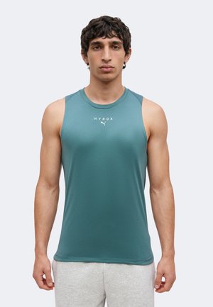 Jeune homme portant un débardeur de sport turquoise avec "HYROX" et le logo Puma, associé à un short gris clair, debout devant un fond uni.