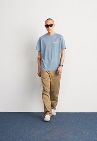 Camiseta de algodón azul claro con un pequeño logo rosa, combinada con pantalones beige y zapatillas blancas. Unas gafas de sol negras completan el look.