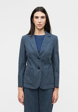 Femme aux longs cheveux foncés portant un blazer bleu marine à fines rayures et un pantalon assorti, debout devant un fond clair uni.