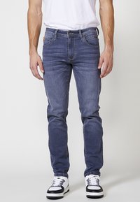 Jeans de mezclilla azul de corte slim con cinco bolsillos, lavado desgastado y detalles de costura. Combinados con zapatillas deportivas blancas y negras.