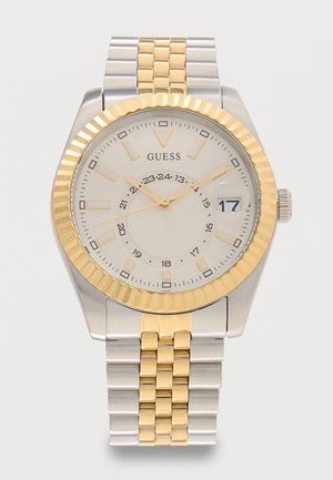Tweekleurige zilver- en goudkleurige metalen polshorloge met witte wijzerplaat, gouden uurmarkeringen en wijzers, datumvenster met de 17 en merknaam Guess.