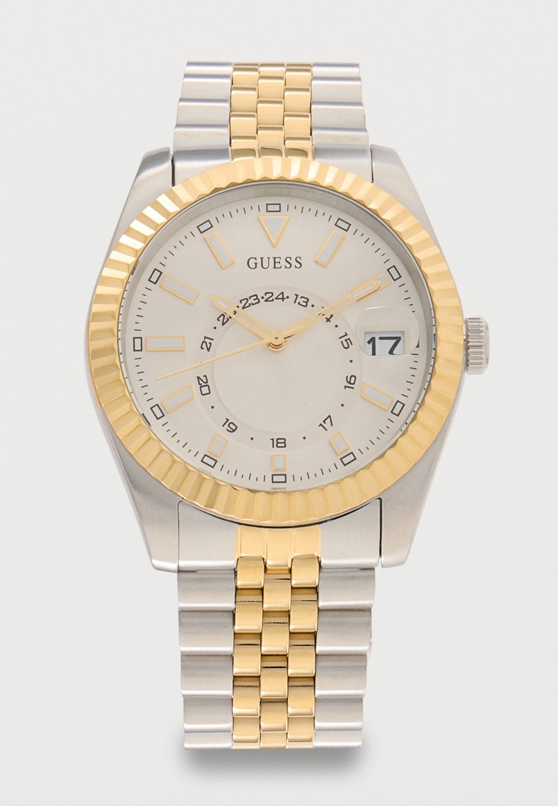 Ανδρικό ρολόι χειρός από ατσάλι δίχρωμο σε ασημί και χρυσό, με λευκό καντράν, χρυσούς δείκτες ώρας και δείκτες, παράθυρο ημερομηνίας που δείχνει 17, και το όνομα του εμπορικού σήματος Guess.