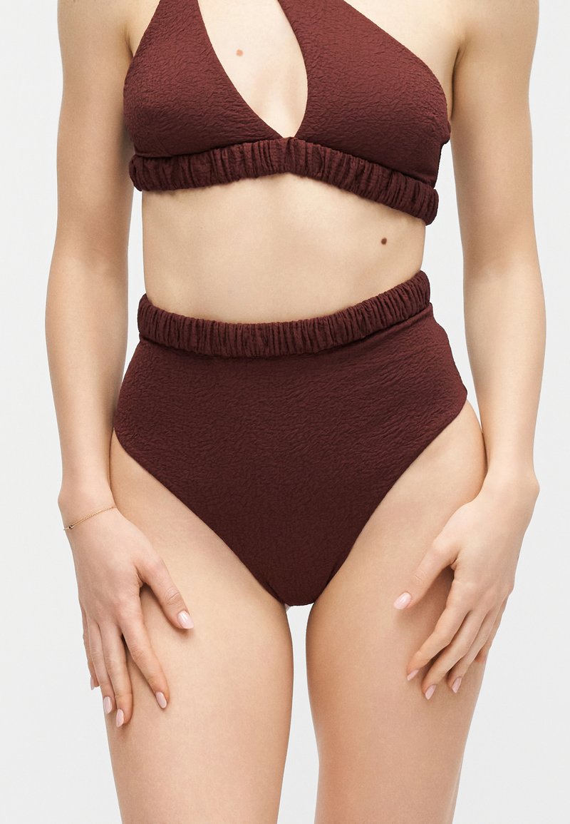 Femme portant un bas de bikini taille haute texturé marron foncé et un haut halter assorti avec des bandes élastiques froncées.