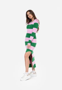 Abito di maglia con strisce orizzontali verdi e rosa, maniche lunghe, scollo rotondo e uno spacco alto fino alla coscia. Abbinato a sneakers bianche.