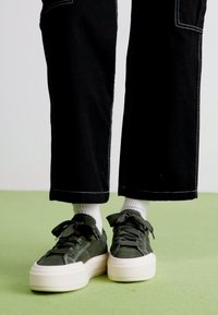 Zapatillas verdes con un upper texturizado, suela de plataforma blanca gruesa y cordones negros, combinadas con calcetines blancos y pantalones negros cortos con costuras blancas.