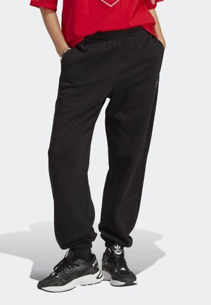 Personne portant un pantalon de jogging noir à taille élastique et des baskets Adidas noires, debout les mains dans les poches devant un fond uni.