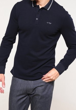 Homme portant un polo bleu marine à manches longues avec col à boutons et un pantalon gris, la main posée dans la poche.