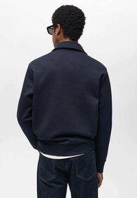 Námornícky sveter so štýlovým golierom, rebrovanými manžetami a lemom. Tkanina vyzerá hladko a má štruktúrovaný strih. Pohľad na chrbát ukazuje minimálne dizajnové detaily.