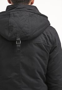 Svart kviltad jacka med huva, som har ett band och spänne bak. Mjuk textur, minimalistisk design och avslappnad silhuett.