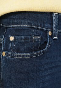 Jean en denim bleu foncé avec une petite poche avant visible, des accents métalliques dorés et des coutures blanches contrastantes le long des coutures.