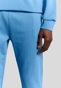 Lichtblauwe joggingbroek gemaakt van zachte stof, met een elastische tailleband en smalle boorden, met een hand die aan de zijkant rust.