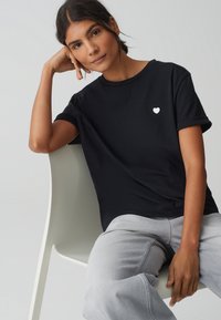 T-shirt nera a maniche corte con un emblema a forma di cuore bianco sul petto, abbinata a jeans grigi chiari, seduta su una sedia bianca.