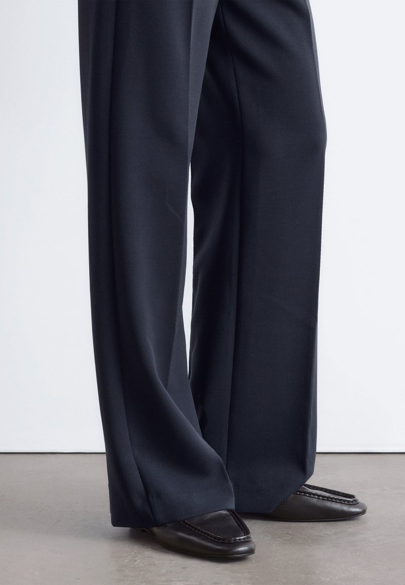 Personne portant un pantalon noir à jambes larges et des mocassins noirs à bout carré, debout sur un sol gris sur fond blanc.