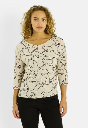 Haut beige à motifs de chats noirs, manches courtes et col rond. Présente un tissu texturé et des boutons accentués sur l'épaule.