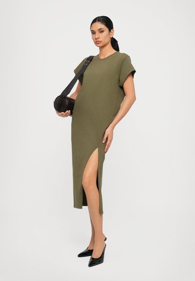 Olive green, knee-length dress with short sleeves and a side slit, paired with a round black bag and pointed black heels.