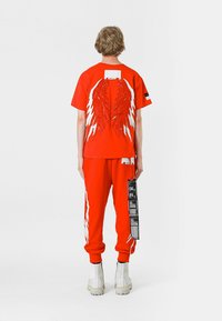 Camiseta corta roja con adornos en forma de alas blancos, combinada con pantalones de chándal rojos a juego con detalles blancos y una etiqueta negra. Botas blancas.