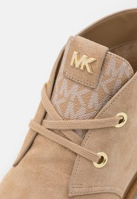 Gros plan sur une chaussure en suède beige avec une languette en tissu de marque affichant un motif répété "MK" et un logo "MK" doré sur le rabat des lacets.