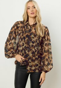Blusa marrone e nera con motivo tie-dye, maniche lunghe a sbuffo e dettagli plissettati. Bottoni frontali e tessuto leggero e trasparente. Abbinata a leggings neri.