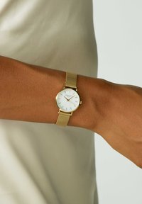 Zlatni mrežasti sat s bijelim brojčanikom, oznakama sati i minimalističkim dizajnom, nosi se na zapešću nasuprot svijetle bež pozadine.