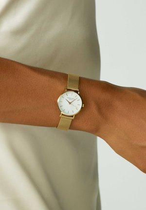 Montre avec un bracelet en maille dorée et un cadran blanc, des index horaires, et un design minimaliste, portée au poignet sur un fond beige clair.