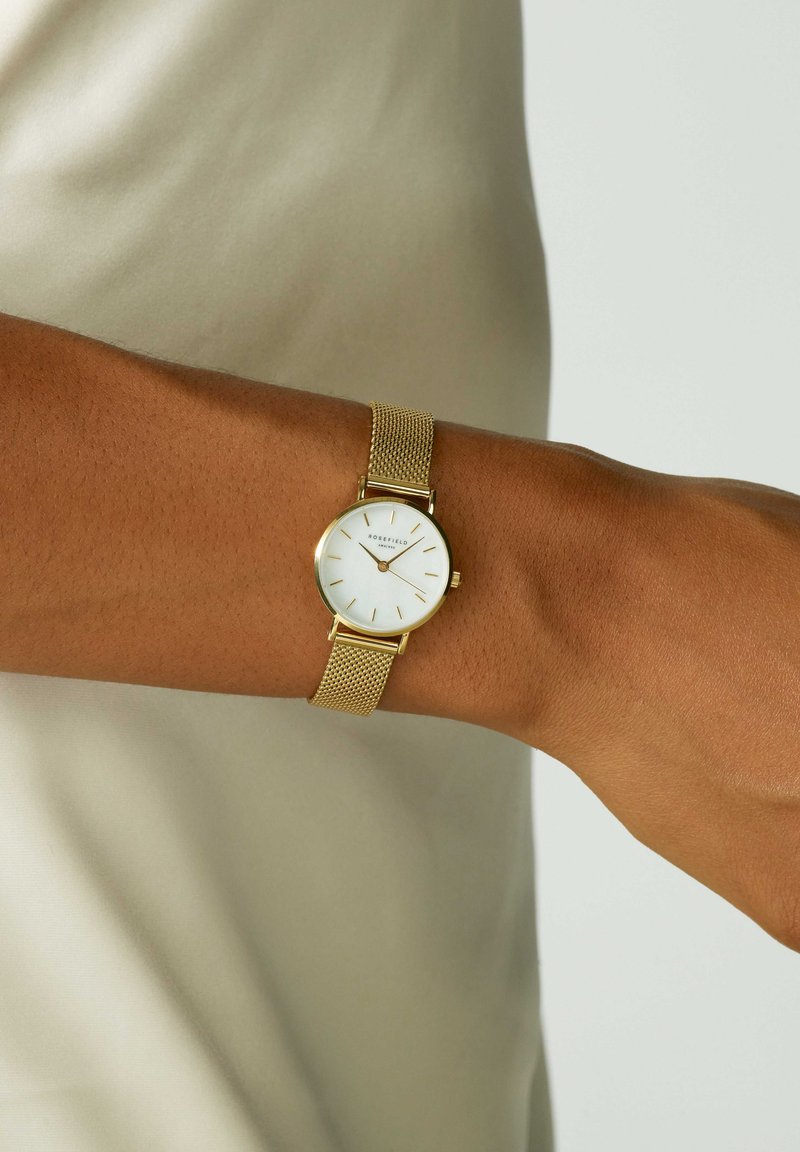 Zlatni mrežasti sat s bijelim brojčanikom, oznakama sati i minimalističkim dizajnom, nosi se na zapešću nasuprot svijetle bež pozadine.