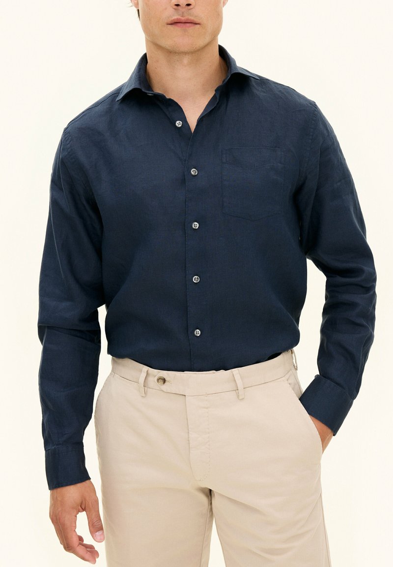 Camicia di lino blu navy con colletto classico, tasca frontale e chiusura con bottoni, abbinata a pantaloni beige chiaro. Tessuto morbido.