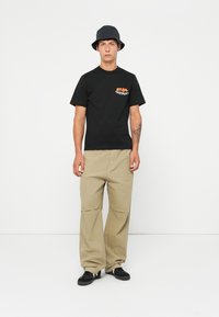 Young man wearing a black bucket hat, black graphic tee, beige wide-leg pants, and black sneakers standing against a white background.