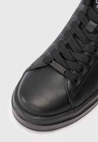 Nahaufnahme eines schwarzen Ledertrainers mit dickem Sohlen und schwarzen Schnürsenkeln, die ein kleines "GUESS"-Tag an den Schnürsenkeln tragen.