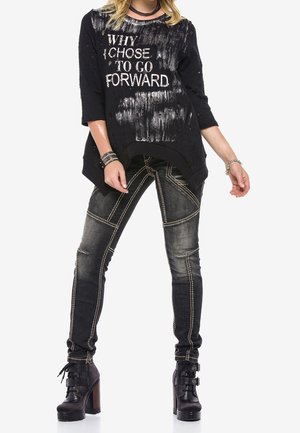 Femme en haut noir déchiré avec le texte "POURQUOI J'AI CHOISI D'AVANCER", jean foncé délavé avec coutures contrastantes, et bottes à talons noires.