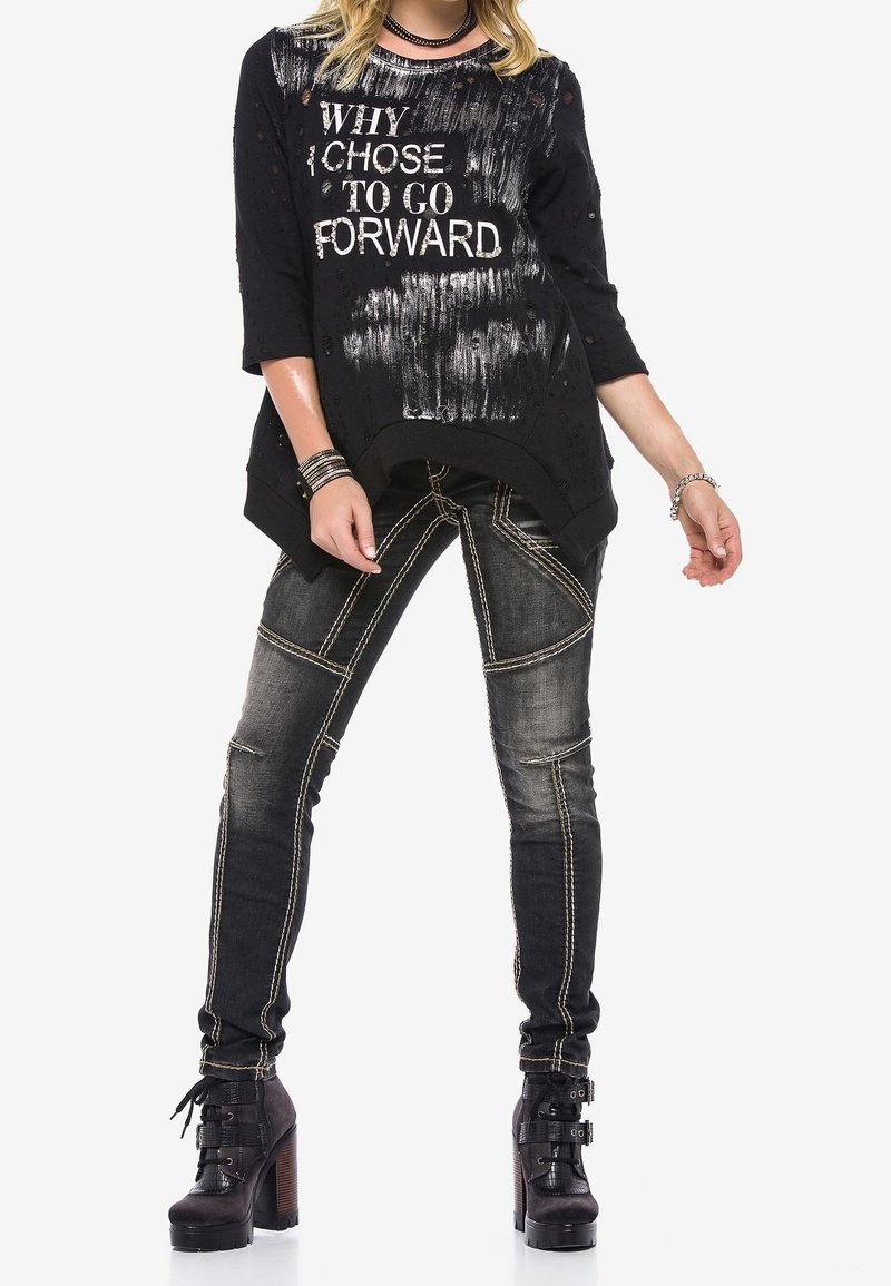 Femme en haut noir déchiré avec le texte "POURQUOI J'AI CHOISI D'AVANCER", jean foncé délavé avec coutures contrastantes, et bottes à talons noires.