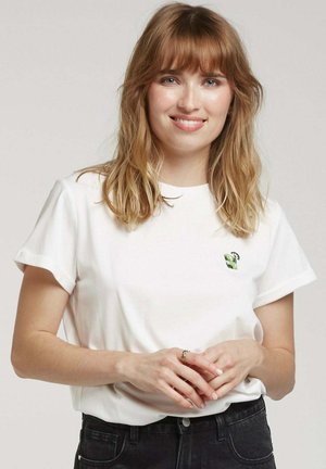 T-shirt blanc à manches courtes retroussées, avec un petit design brodé vert et noir sur la zone gauche de la poitrine. Jeans noirs visibles.