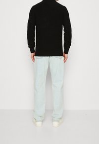 Hombre con un suéter negro de manga larga, jeans sueltos de color azul claro y zapatillas blancas, de pie frente a una pared blanca.