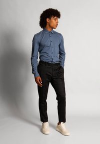 Hemd zum Knöpfen in Hellblau, kombiniert mit dunkel karierten Hosen. Schuhe sind cremeweiße Sneakers. Das Model steht im Profil vor einem einfarbigen Hintergrund.