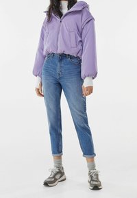 Fialová péřová bunda s kapucí, elastickými manžetami a přední kapsou, v kombinaci s vysokým pasem modrými džíny a šedými teniskami s texturovanou podrážkou.