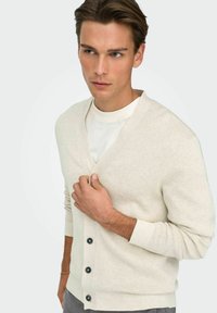 Beige cardigan van zachte gebreide stof, met een V-hals, lange mouwen en vijf donkere knopen aan de voorkant. Gladde textuur en een comfortabele, losse pasvorm.