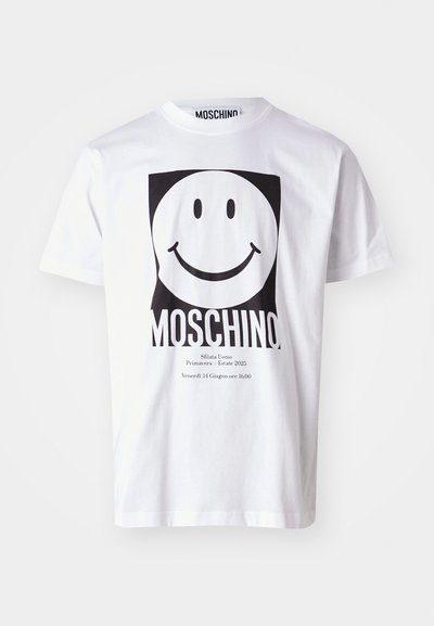 Balts kokvilnas T-krekls ar lielu melnu smaidošu seju grafiku, ar "MOSCHINO" izdrukātu zemāk un pasākuma detaļām mazākā tekstā.