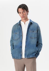 Denim košulja-jakna u srednje plavoj boji, s prednjim dugmadi, dva džepa na prsima i ležernim krojem. Nosi se preko bijelog polo majice i bež hlača.