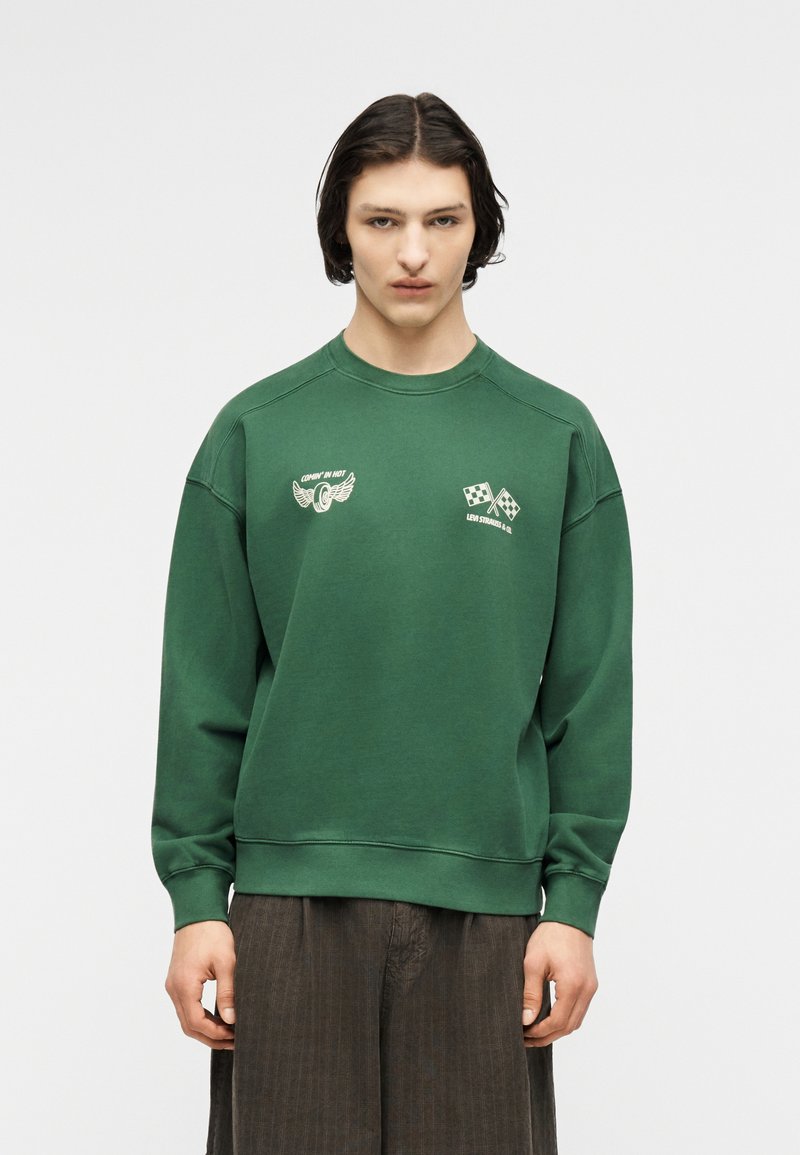 Jeune personne aux cheveux mi-longs foncés, portant un sweat-shirt vert avec des logos de drapeau de course blanc et de roue ailée, et un pantalon foncé, immobile.