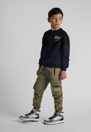 Schwarzes Sweatshirt mit Text, grüne Cargo-Hose mit Taschen und Patches sowie schwarz-weiße Sneaker. Das Modell steht mit einem neutralen Gesichtsausdruck.