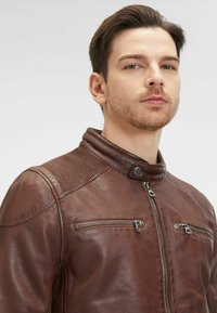 Giubbotto in pelle marrone con colletto alto, tasche con zip e accenti a coste sulle spalle. Texture liscia e design aderente.