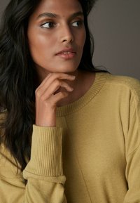 Pull en maille jaune moutarde avec un col et des poignets côtelés. Texture lisse, coupe décontractée, et présentant un détail de couture sur le côté.