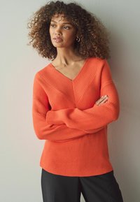 Ribbad orange tröja med V-ringning och diagonala sömmar, med långa ärmar och en avslappnad passform, tillsammans med svarta byxor.