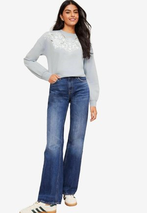 Lys blå sweatshirt med blonde detaljer på brystet, parret med mørkeblå flared jeans og hvide sneakers med grønne accenter.