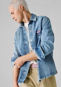 Person med kort blondt hår ruller opp ermene på en blå denim Red Bull Racing-jakke over hvit skjorte og beige bukser.