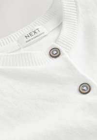 Pull blanc avec un col côtelé et deux boutons marron. L'étiquette indique "NEXT" avec "FABRIQUÉ AU BANGLADESH" en dessous. La texture douce est visible.